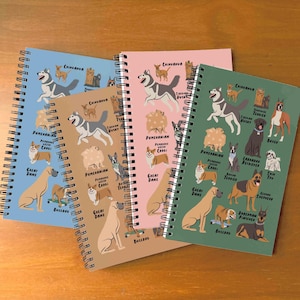Puede incluir: Tres cuadernos de espiral con diferentes razas de perros ilustradas en las portadas. Las portadas son marrones, rosas y verdes. Cada cuaderno presenta una raza de perro diferente en cada página, con el nombre de la raza impreso debajo de cada perro.