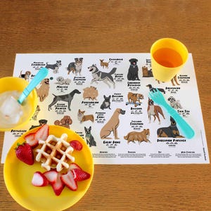 Puede incluir: Un mantel individual blanco con ilustraciones de diferentes razas de perros, incluyendo un Rottweiler, un Braco Alemán de Pelo Corto, un Pomerania, un Gran Danés, un Shetland Sheepdog y un Doberman Pinscher. El mantel individual está sobre una mesa de madera con un tazón amarillo, una taza amarilla y un plato amarillo con fresas y un waffle.