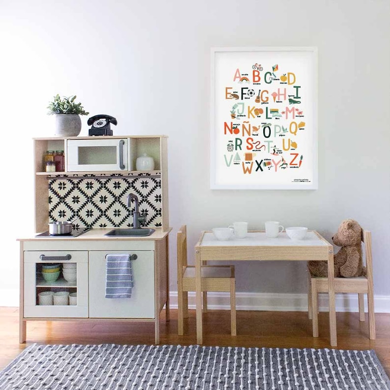 May include: A white play kitchen with a black and white patterned backsplash, a microwave, and a sink. A wooden table and chairs are set up in front of the kitchen. A framed alphabet poster with colorful illustrations hangs on the wall.