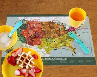 Puede incluir: Un mantel individual verde y blanco con un mapa de los Estados Unidos, que muestra los estados y los parques nacionales. El mantel individual está sobre una mesa de madera con un tazón amarillo, un plato amarillo con fresas y un waffle, una taza amarilla y un tenedor azul.