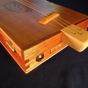 Puede incluir: Una guitarra de caja de puros de madera con un cuerpo de madera clara y un cuello de madera oscura. La guitarra tiene una sola pastilla y un puente con tres clavijas de afinación. La guitarra está hecha de una caja de puros reutilizada.