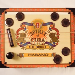 Puede incluir: Una guitarra hecha a mano con una caja de cigarros. El cuerpo crema presenta el texto "Spirit of Cuba" y "Alec Bradley Habano". Tiene un mástil de madera, cuerdas y clavijas negras. Un instrumento musical único.