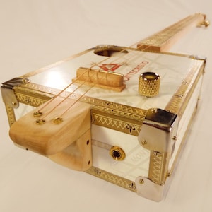 May include: A white cigar box guitar with gold trim and a wooden neck. The guitar has two strings and a gold tuning knob. The box has the text "Montecristo" printed on it.
