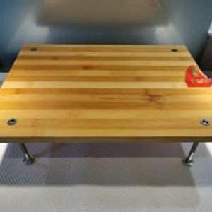 Könnte beinhalten: Ein rechteckiges Bambus-Schneidebrett mit einem roten, eckigen Plastikschutz. Das Brett hat vier Metallbeine und steht auf einer weißen Oberfläche.