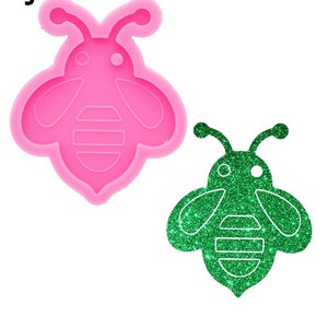 May include: Pink silicone mould of a bee with a green glitter bee in the mould. The bee has a simple design with wings, a body, and a head with two eyes.