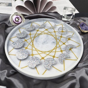 Puede incluir: Una bandeja de mármol blanco con un diseño geométrico dorado y ocho fases lunares de purpurina plateada.