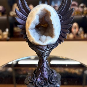Peut inclure: Un œuf en géode d'agate blanche avec un centre cristallin creux, maintenu par un support métallique violet foncé. Le support présente des ailes sculptées s'étendant vers le haut et une base décorée de formes florales en couches.