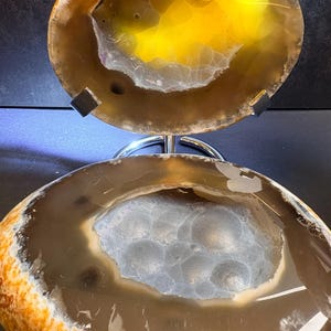 May include: A polished agate geode slice featuring a translucent brown outer ring and a central cavity filled with sparkling, light-coloured crystalline formations, displayed on a silver-toned metal stand that holds the stone in an upright position.