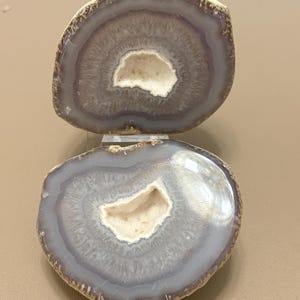 Peut inclure: Une géode d'agate fendue présentant des bandes concentriques de couches minérales gris clair et blanches translucides. Le centre de chaque moitié révèle une cavité creuse tapissée de petits cristaux de quartz blancs étincelants, entourée d'une croûte extérieure brune et rugueuse.