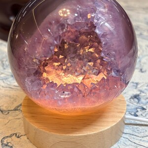 May include: A spherical amethyst geode lamp featuring a polished outer surface and a hollow interior filled with jagged purple crystals. The geode is illuminated from within by a warm light and rests on a circular light-colored wooden base.