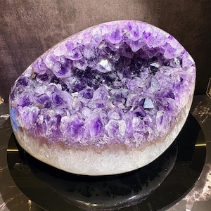 Pode incluir: Uma grande geoda de ametista arredondada com pontas cristalinas roxas profundas agrupadas dentro de uma cavidade oca em forma de tigela. A base externa do espécime mineral exibe uma formação rochosa branca, áspera e translúcida.