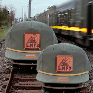 May include: Two olive green baseball caps with a brown leather patch featuring a train illustration and the letters "BMFS." Each cap has a yellow rope detail and a small orange label. A blurred train is in the background.