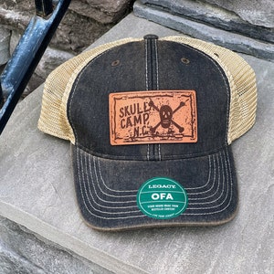 May include: A black and tan trucker hat with a brown leather patch that says "Skull Camp NC" with a skull and crossbones. The hat also has a green circular patch that says "Legacy OFA" with the text "Hand Crafted Made from Recycled Content Live Your Legacy".