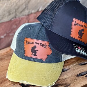 May include: Two black and grey trucker hats with a brown leather patch that says "Iowans For Panic" and a silhouette of a person sitting in a chair. The patch is shaped like the state of Iowa.