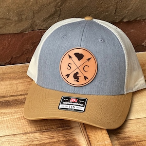 May include: A gray and brown baseball cap with a white mesh back. The cap features a brown leather patch with a silhouette of South Carolina and the letters "S" and "C" with arrows crossing in the middle. The patch is attached to the front of the cap.