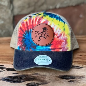 May include: A tie-dye trucker hat with a brown leather patch featuring a dancing figure. The hat has a black brim and a mesh back. The patch has the text "OLD FAVORITE STRUCTURED" and "LEGACY" on it.