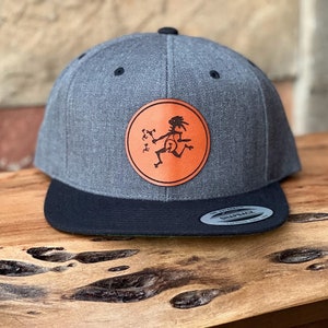 May include: A gray and black baseball cap with a flat brim. The cap features a circular orange patch with a black graphic of a running figure. The cap is sitting on a wooden surface.