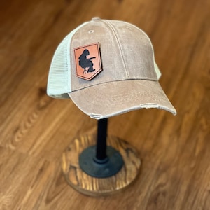 May include: A brown and white trucker hat with a distressed look. The hat has a brown leather patch with a black silhouette of a person sitting on a chair.