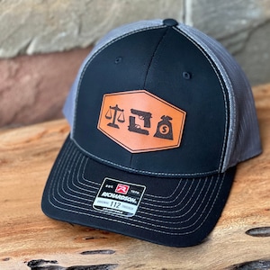 May include: Black and gray trucker hat with a brown leather patch featuring a scale, hammer, and money bag. The patch has the text "JUSTICE" on it. The hat is adjustable and has the Richardson 112 logo on the back.