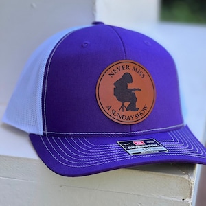 May include: Purple and white trucker hat with a brown leather patch that says "Never Miss A Sunday Show" and a silhouette of a person playing a guitar. The hat is made by Richardson 112.