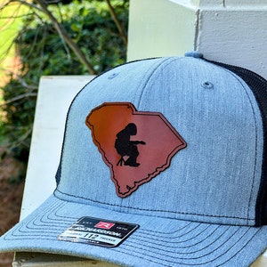 May include: A blue and black trucker hat with a brown leather patch featuring the outline of South Carolina and a silhouette of a person sitting on a stool. The hat has a black mesh back and a Richardson 112 label.