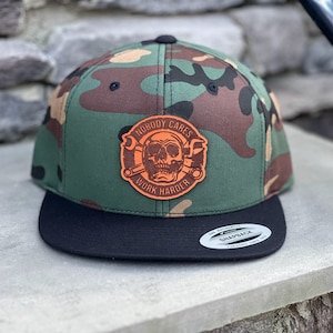 May include: A camouflage green and brown baseball cap with a black brim. The cap has a brown leather patch with a skull and crossbones design and the text "Nobody Cares Work Harder".