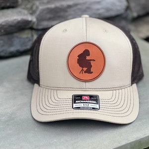 May include: A tan and black trucker hat with a brown leather patch featuring a silhouette of a person sitting on a stool. The patch is circular and has a red border. The hat has a black mesh back and a brown adjustable strap with the text "RICHARDSON. EST. 1970 ORIGINAL 112 TRUCKER ADJUSTABLE".