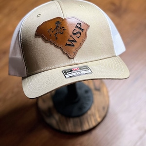 May include: A beige and white trucker hat with a brown leather patch shaped like the state of South Carolina. The patch features a stick figure design and the text "WSP".