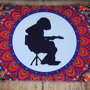 May include: A black silhouette of a person playing a guitar, sitting on a stool, inside a white circle with a colorful mandala pattern background.