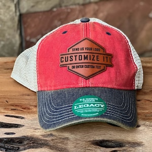 May include: A red, navy blue, and tan trucker hat with the text "CUSTOMIZE IT!" on a leather patch. The hat has a mesh back and a curved brim. The hat also has a green and white sticker that says "LEGACY".