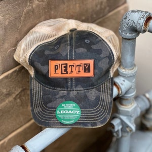 May include: A black and camo trucker hat with a brown leather patch that says "PETTY" in white letters. The hat also has a green circular patch that says "OLD FAVORITE TRUCKER LEGACY" in white letters.