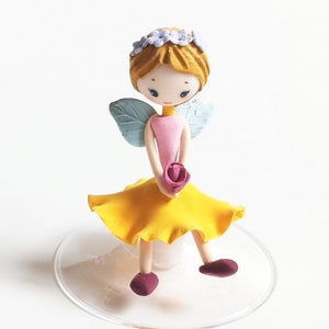Könnte beinhalten: Eine kleine, gelbrockige Feenfigur mit blauen Flügeln, einem rosa Kleid und einer lila Blume. Sie trägt eine lila Blütenkrone und hat blondes Haar.