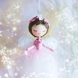 以下が含まれることがあります： 目を閉じ、ピンクのドレスと花冠をつけたピンクと白の妖精人形。人形は白いチュールスカートとキラキラした背景に囲まれています。