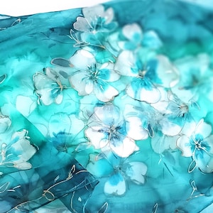May include: A teal and turquoise silk scarf with a floral pattern. The scarf features white and light blue flowers with gold accents. The fabric has a watercolor effect, with soft gradients of color.