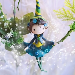 Peut inclure: Une figurine de fée fantaisiste avec un chapeau vert et jaune, une robe bleue et des accents de fleurs bleues. La fée est perchée sur une branche avec un fond enneigé.