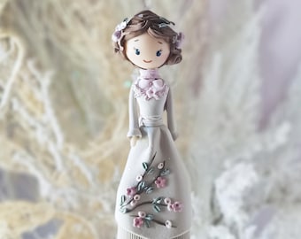 May include: A decorative figurine featuring a person with brown hair adorned with small light purple flowers. The figure wears a long, light grey dress with a high collar, a pink neck accent, and a floral pattern of pink blossoms and green leaves.
