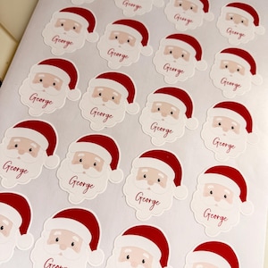 May include: A sheet of twenty individual stickers, each featuring a cartoon Santa Claus face wearing a red hat. Below each Santa face, the name George is printed in a red cursive script. The stickers are arranged in a grid pattern.