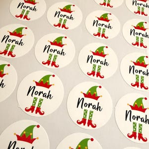 May include: A sheet of circular white stickers, each featuring the name Norah in black script, topped with a green and red elf hat and finished with green and red elf legs with curled toes at the bottom of the text.