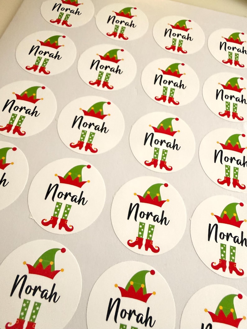 May include: A sheet of circular white stickers, each featuring the name Norah in black script, topped with a green and red elf hat and finished with green and red elf legs with curled toes at the bottom of the text.