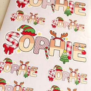 May include: A collection of die-cut stickers featuring the name SOPHIE in a festive Christmas theme. Each letter is decorated with red and white plaid patterns, elf hats, reindeer antlers, candy canes, Christmas trees, and string lights.