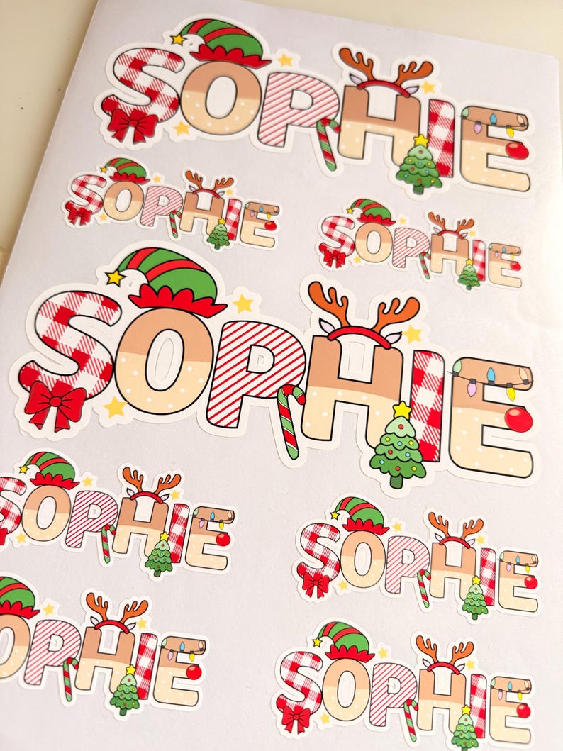 May include: A collection of die-cut stickers featuring the name SOPHIE in a festive Christmas theme. Each letter is decorated with red and white plaid patterns, elf hats, reindeer antlers, candy canes, Christmas trees, and string lights.