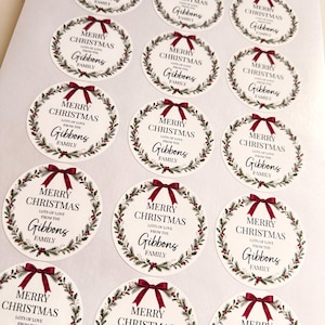May include: A sheet of circular stickers featuring a festive wreath design with a dark red bow at the top. The text reads Merry Christmas, lots of love from the Gibbons family, printed in black ink on a white background.