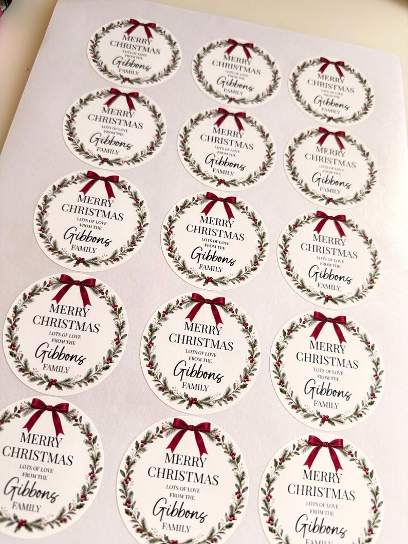 May include: A sheet of circular stickers featuring a festive wreath design with a dark red bow at the top. The text reads Merry Christmas, lots of love from the Gibbons family, printed in black ink on a white background.