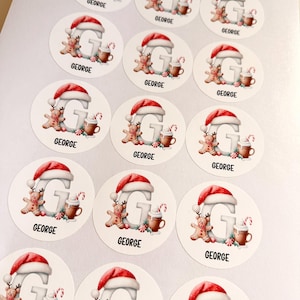 May include: A sheet of circular stickers featuring a large letter G wearing a Santa hat, accompanied by a gingerbread person, a mug of hot cocoa, and a candy cane. Each sticker includes the name GEORGE printed in black capital letters.