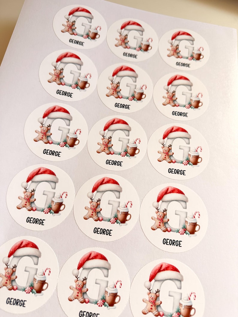 May include: A sheet of circular stickers featuring a large letter G wearing a Santa hat, accompanied by a gingerbread person, a mug of hot cocoa, and a candy cane. Each sticker includes the name GEORGE printed in black capital letters.