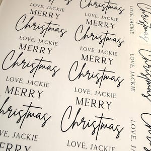 May include: A sheet of white adhesive labels featuring black printed text. Each label displays the words MERRY Christmas in a combination of serif and script fonts, followed by the text LOVE, JACKIE in a simple sans-serif typeface.