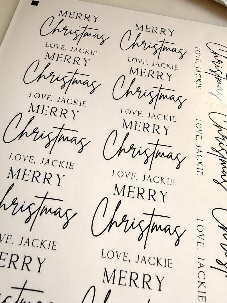 May include: A sheet of white adhesive labels featuring black printed text. Each label displays the words MERRY Christmas in a combination of serif and script fonts, followed by the text LOVE, JACKIE in a simple sans-serif typeface.