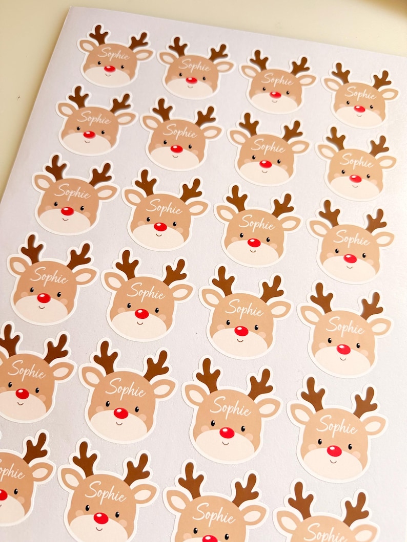 May include: A sheet of circular stickers featuring a cartoon reindeer face with brown antlers, a red nose, and the name Sophie written in white cursive script across the forehead of each individual reindeer illustration.