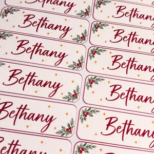 May include: A sheet of rectangular adhesive labels featuring the name Bethany in a red script font. Each label is decorated with green holly leaves, red berries, and small gold stars on a cream background, arranged in two vertical columns.