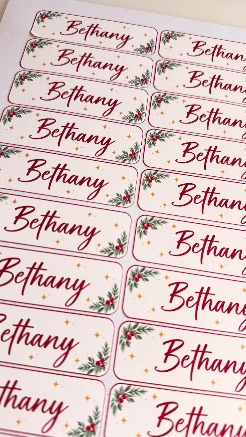 May include: A sheet of rectangular adhesive labels featuring the name Bethany in a red script font. Each label is decorated with green holly leaves, red berries, and small gold stars on a cream background, arranged in two vertical columns.